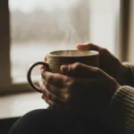 A close-up, cinematic shot of a person's hands wrapped gently around a warm, textured ceramic mug of coffee on a rainy morning. Soft, natural light filters through a nearby window, illuminating the faint steam rising from the mug. The mood is tranquil, safe, and deeply grounding, capturing the essence of a micro-moment of peace. High resolution, photorealistic, soft focus background.