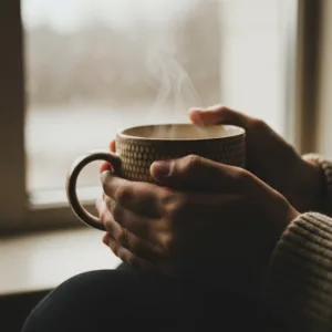 A close-up, cinematic shot of a person's hands wrapped gently around a warm, textured ceramic mug of coffee on a rainy morning. Soft, natural light filters through a nearby window, illuminating the faint steam rising from the mug. The mood is tranquil, safe, and deeply grounding, capturing the essence of a micro-moment of peace. High resolution, photorealistic, soft focus background.