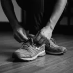 A cinematic, close-up shot of a person's hands tightly lacing up worn running shoes on a dimly lit wooden floor. The lighting is moody and warm, capturing the quiet, unglamorous moment of taking the very first step before motivation arrives.