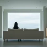 A serene, minimalist living room with a single person sitting comfortably on a modern sofa, looking out a large window at a quiet, overcast sky. No electronics or books are visible. Soft, natural, diffused lighting, cinematic photography, high resolution.