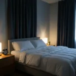 A peaceful, dimly lit bedroom optimized for sleep, featuring a neatly made bed with breathable linens, blackout curtains drawn, and a small analog alarm clock on the wooden nightstand. Warm, low-intensity ambient lighting.