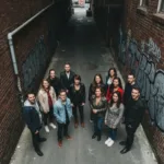 A conceptual representation of anticipatory anxiety in a workspace. A person sits at a clean, minimalist wooden desk, but faint, glowing silhouettes of floating notification icons, envelopes, and ticking clocks hover in the dark, moody background, illustrating the invisible weight of expected interruptions. Cinematic lighting, photorealistic.