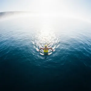 A lone swimmer in the middle of a vast, calm lake, viewed from above. The starting shore and the destination shore are both barely visible in the misty distance. The water is deep blue, conveying a sense of scale and isolation, illustrating the psychological 'messy middle' of a long journey. Cinematic lighting, hyper-realistic.