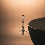 A cinematic, moody photograph of a single drop of water overflowing from the rim of a simple, elegant ceramic cup, capturing the exact moment the surface tension breaks. Soft, diffused lighting, neutral minimalist background, emphasizing the psychological concept of capacity and overwhelm.