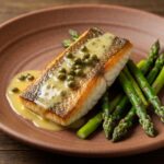 A beautifully plated pan-seared barramundi fillet with crispy golden skin, topped with a glossy lemon-caper butter sauce, served on a rustic ceramic plate next to vibrant green roasted asparagus. High-end food photography, natural studio lighting, macro detail.