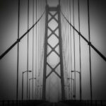 A dramatic, high-contrast black and white photograph of a massive suspension bridge emerging from thick fog. The thick steel cables are taut, emphasizing extreme tension and structural integrity. Cinematic lighting, architectural focus, highly detailed, 8k resolution.
