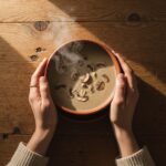 A top-down view of a person's hands wrapped gently around a warm, steaming bowl of soup on a rustic wooden table. Soft, warm natural lighting highlights the rising steam, conveying a deep sense of emotional warmth, grounding, and psychological safety.