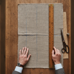 Overhead macro shot of a bespoke tailor measuring a pressed woven linen test swatch with a vintage wooden meter stick on an oak cutting table, showing clear basted chalk marks and fine fabric texture