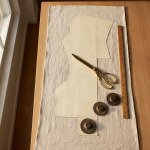 A clean overhead flat lay of a tailor's oak cutting table with raw linen fabric, sharp brass tailoring shears, a wooden meter stick, and paper pattern pieces carefully nested with weights, soft natural atelier morning light.